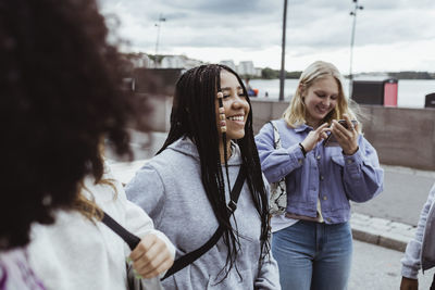 Teenage girl using smart phone standing with female friends on footpath