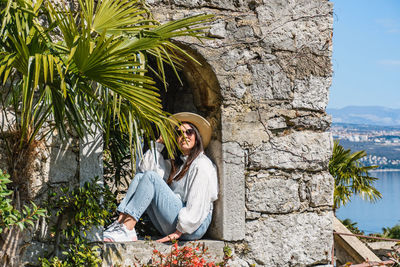 Attractive young woman in spring outfit sitting in window of stone building in park.