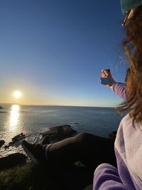 Woman looking into small mirror against sky during sunset