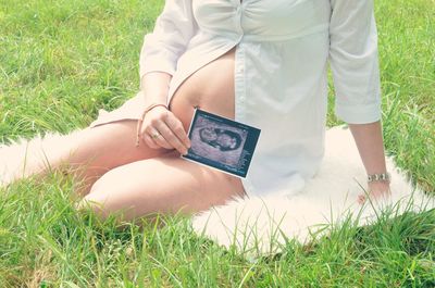 Midsection of pregnant woman holding ultrasound photograph while sitting on grassy field