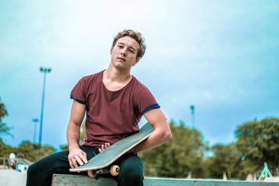 Portrait of young man with skateboard sitting against sky