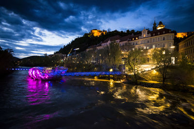 Illuminated city by river against sky at dusk