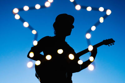 Low angle view of silhouette man playing guitar by illuminated lighting equipment against sky