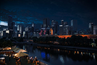 Illuminated buildings in front of river against sky at night