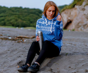 Young woman holding insulated drink container while sitting at beach