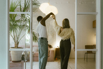 Carefree young man in undershirt holding hands while dancing with girlfriend at home