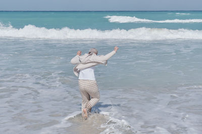 Senior caucasian woman with gray hair jumping out of incoming wave in sandy beach of atlantic ocean