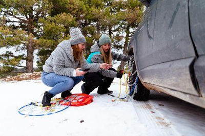 Female friends in warm activewear looking at instruction on smartphone and at manual guide trying to put security snow chain on wheel tire while squatting near stuck car in snowy road