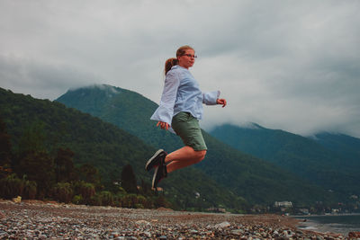 Low angle view of woman jumping at beach against cloudy sky