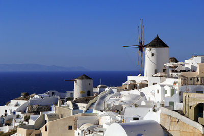 Traditional windmills amidst buildings on mountain by sea against clear blue sky