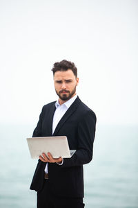 Businessman using laptop while standing at beach against clear sky