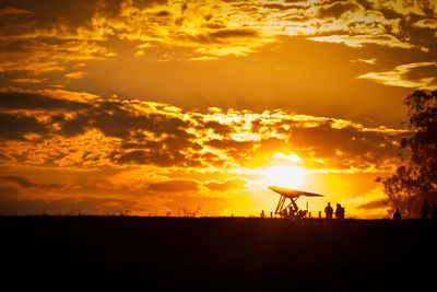 People preparing for hang-gliding on silhouette field against dramatic sky during sunset