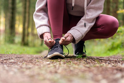 Cropped unrecognizable portswoman in activewear tying shoelaces on sneakers during workout in woods while having break on path