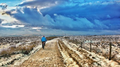 Rear view of man riding bicycle on field against cloudy sky