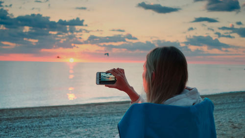 Side view of woman photographing sea against sky during sunset