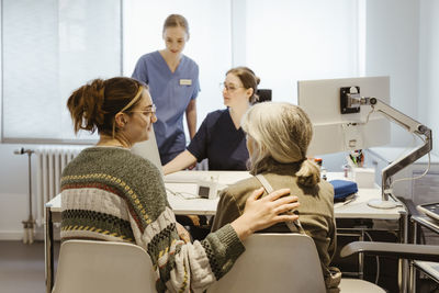 Daughter with hand on shoulder of senior mother sitting on chair during medical exam at hospital