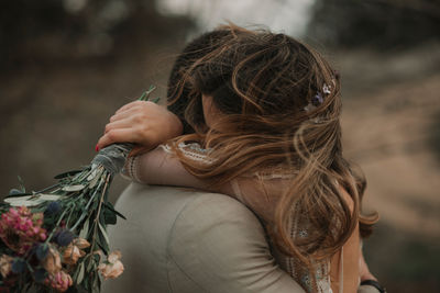 Side view of young guy in suit embracing with lady in wedding dress with bunch of flowers in hand near trees on blurred background