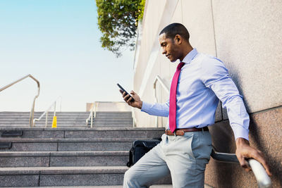 Young man using mobile phone while standing against wall