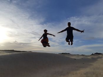 Full length of couple jumping on sand dune against sky