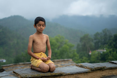 A young indian boy in yellow dhoti meditating in the mountains, with fog.
