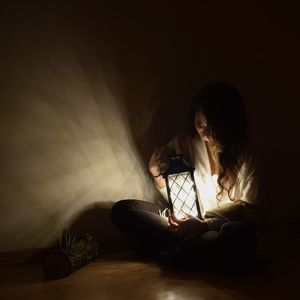 Woman holding illuminated lamp while sitting on bed at home