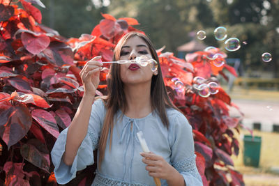 Beautiful young woman blowing bubbles while standing against plants