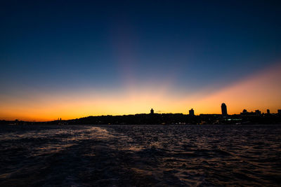 Silhouette city by sea against sky at sunset