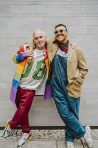 Gay friends with arms around standing on footpath against wall