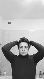 Portrait of serious woman with hands behind head standing in bathroom