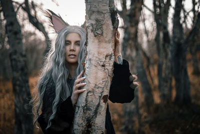Portrait of young woman standing by tree trunk in forest