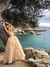 Female model wearing evening gown while leaning on tree trunk at beach