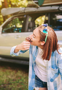 Young woman eating ice cream cone while standing against car