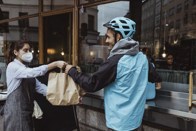 Female owner giving package order to delivery man outside deli shop during pandemic