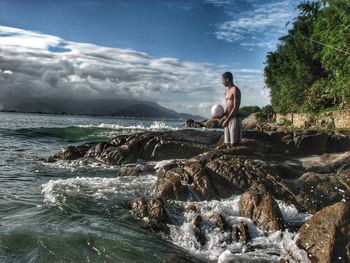 Shirtless man holding ball while standing at rocky beach