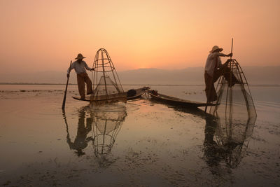 Fishermen fishing in lake against sky during sunset