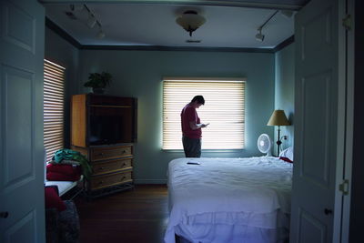 Woman using smart phone while standing against window in bedroom