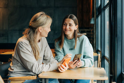 Two woman friends sitting in a cafe watch online media in the phone and laugh.