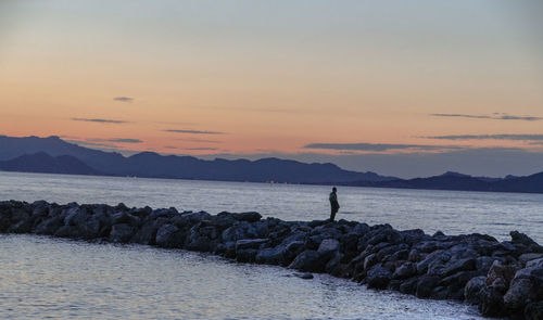 Man standing on groyne at beach against sky during sunset