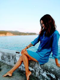 Woman sitting on retaining wall by sea against clear blue sky