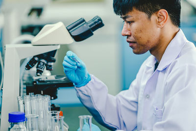 Asian man lab technician in protective glasses and gloves sits next to a microscope in laboratory.