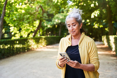 Positive elderly female athlete with gray hair smiling and reading message in social media on smartphone while standing on park alley during break in fitness training