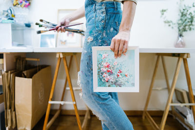 Side view of anonymous female artist standing near easel and holding a colorful paint and brushes in creative workshop