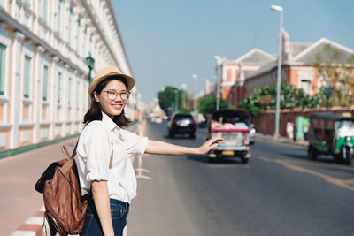 Young asian woman with backpack and straw hat hailing a taxi or tuk tuk in bangkok, thailand