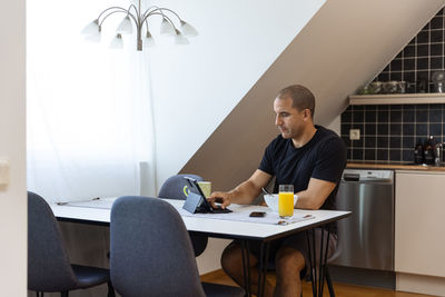 Focused male surfing internet on tablet while sitting at table at home and enjoying breakfast in morning