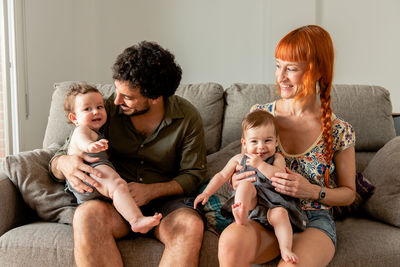 Happy multiethnic young mother and father hugging cute smiling twin babies while sitting on comfortable sofa during weekend at home