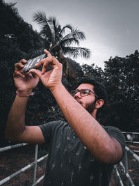 Full length portrait of young man holding sunglasses against trees