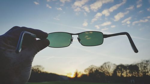 Cropped hand holding sunglasses against sky during sunset