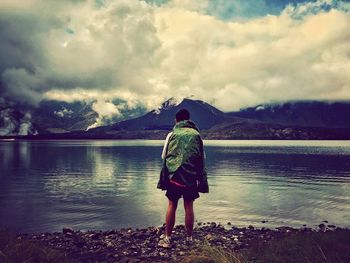 Rear view of man standing at shore against cloudy sky