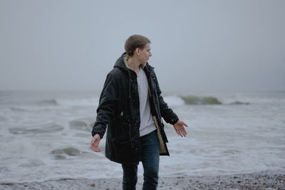 A guy in a warm winter jacket stands on the seashore, spreads his arms and looks to the side