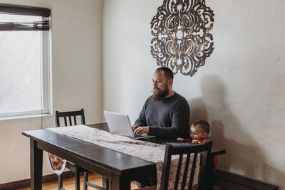 Wide view of father trying to work from home with toddler standing